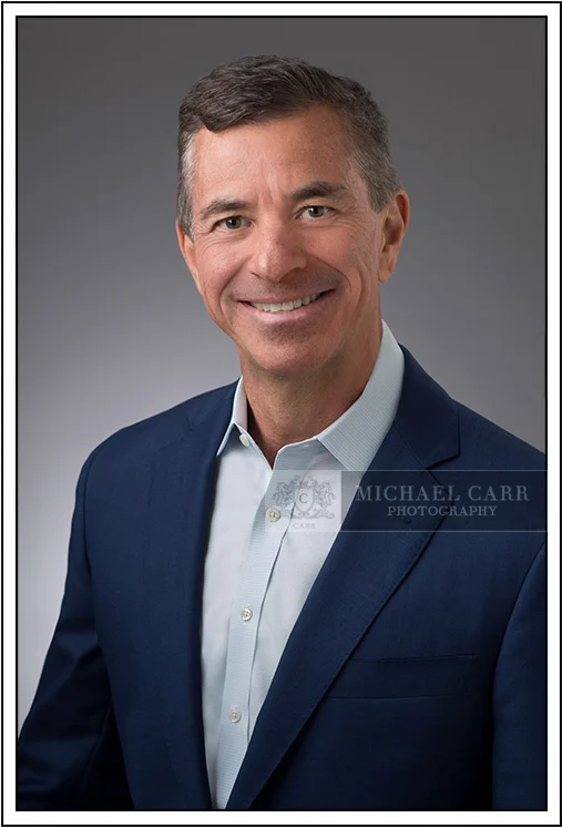 Houston Headshot Photographer Classic executive business headshot of a man wearing a blue suit jacket and light blue dress shirt on a gray studio background in Houston, Texas.